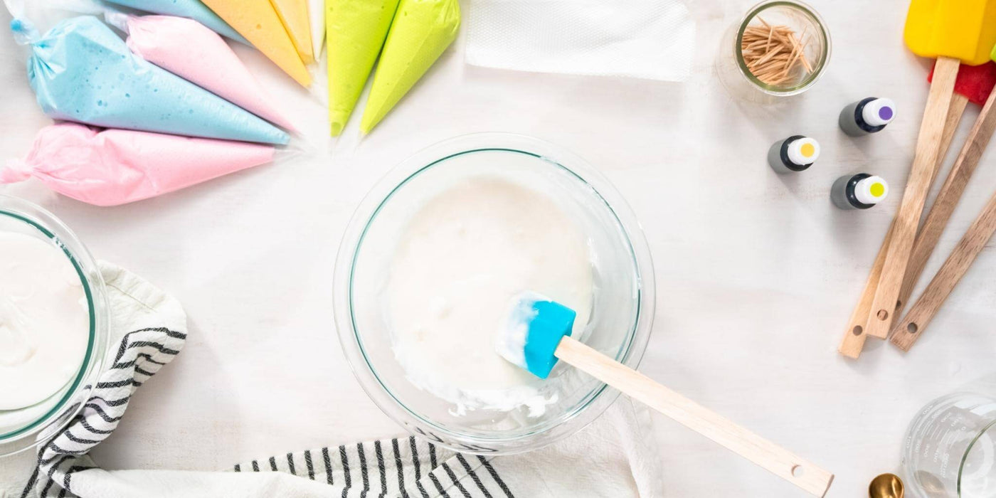 An image of pastel colored icing bags, a mixing bowl with icing in it, food coloring vials, spatulas and a kitchen towel, all on a white marble surface.