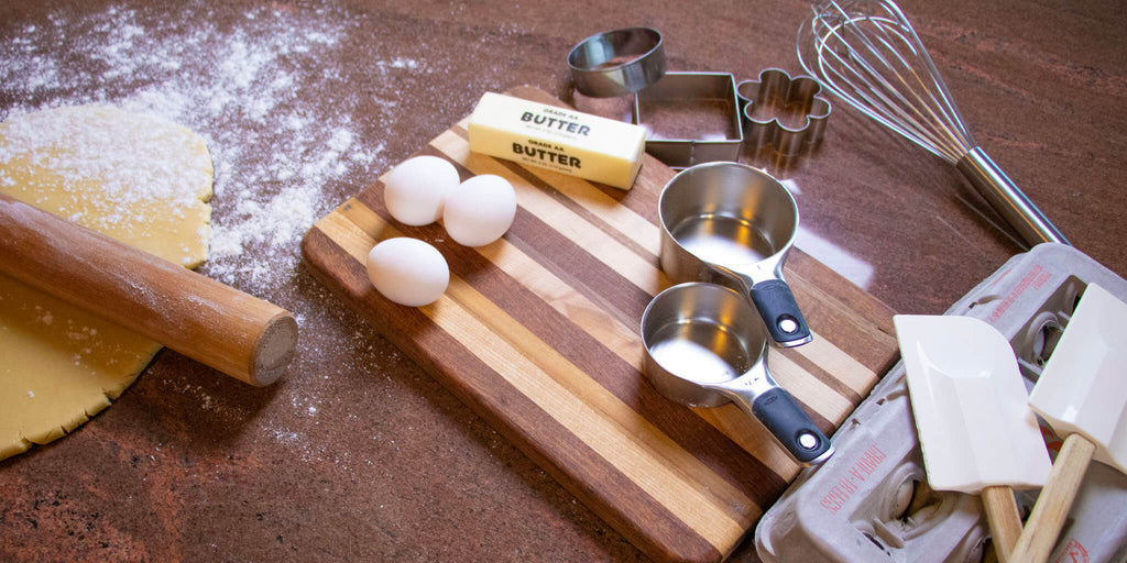 Image of a rolling pin on cookie dough, a cutting board with eggs, measuring cups and cookie cutters on top, a whisk, spatulas and egg carton to the side.